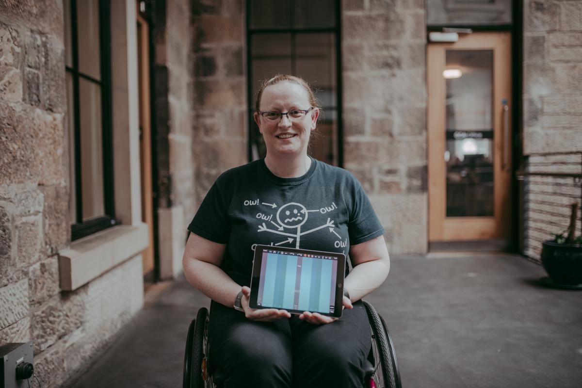 Image of a lady with long blonde hair in a manual wheelchair holding up an iPad displaying a musical keyboard